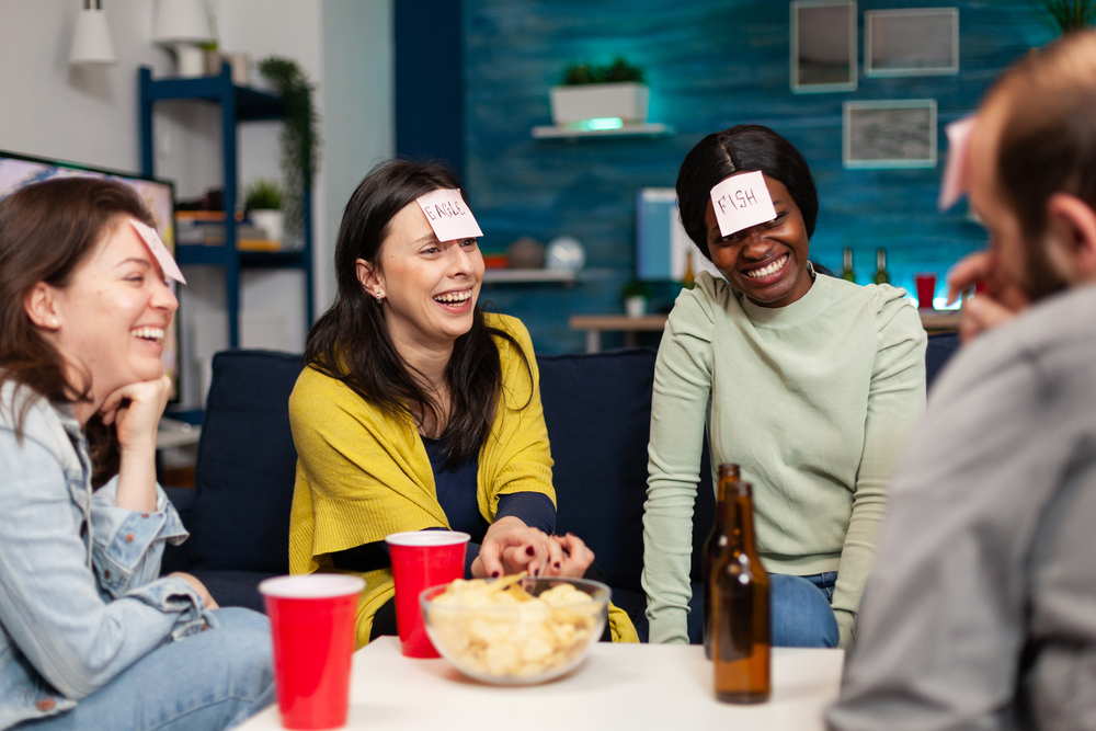 Mixed race friends gathering in living room playing guess who game using sticky notes. Mixed race people having fun, laughing together while sitting on sofa late at night during funny party.