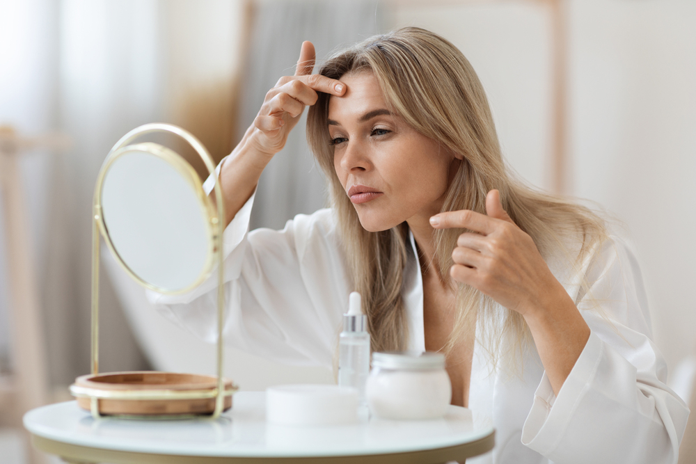 Blonde middle aged woman in white bathrobe sitting at table full of cosmetics, looking at mirror. checking her forehead for pimples and wrinkles, side view. Skin problems concept