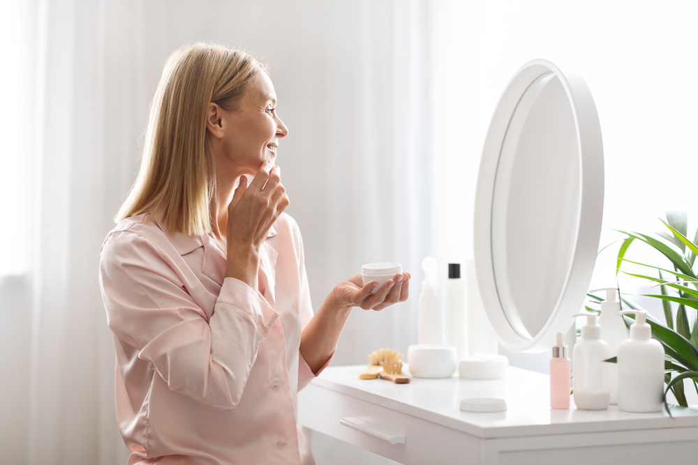 Skincare Concept. Attractive Middle Aged Lady Applying Moisturising Face Cream At Home, Smiling Beautiful Mature Woman Making Beauty Routine While Sitting At Dressing Table In Bedroom