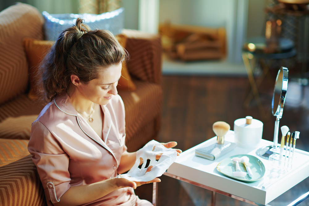 smiling elegant woman in pajamas with no makeup looking at white sheet facial mask at modern home in sunny winter day.