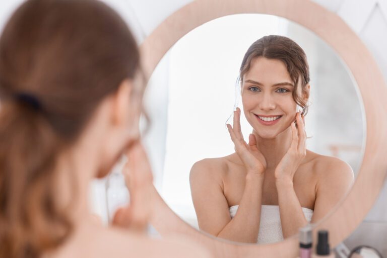 Happy girl enjoying her young smooth skin, looking at mirror at home